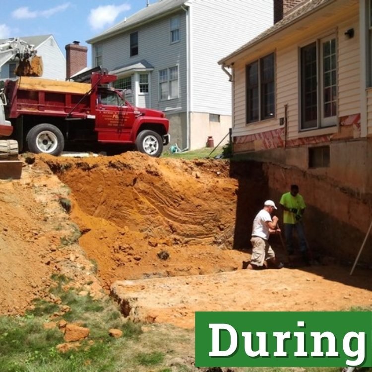 two men dig in a ditch with a red construction truck in the background